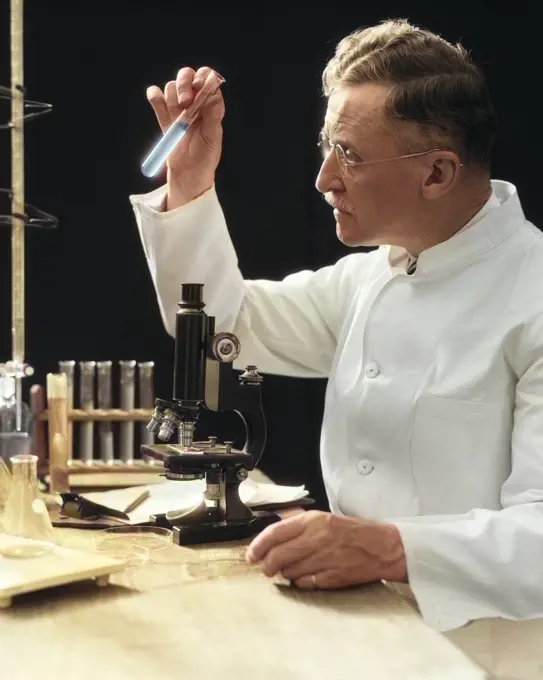1920s 1930s 1940s MAN SCIENTIST LAB TECHNICIAN IN WHITE COAT LOOKING AT TEST-TUBE IN FRONT OF MICROSCOPE