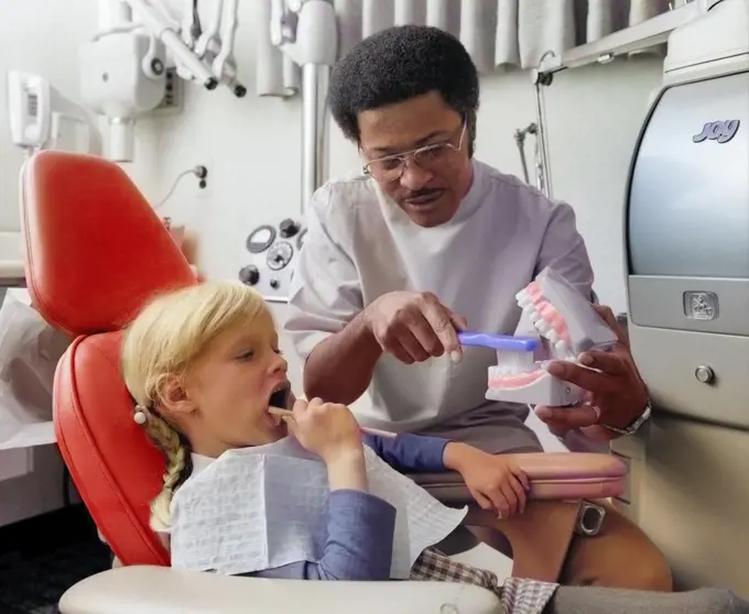 1970s AFRICAN AMERICAN MAN DENTIST TEACHING LITTLE BLOND CAUCASIAN GIRL HOW TO BRUSH HER TEETH USING LARGE MODEL OF JAW