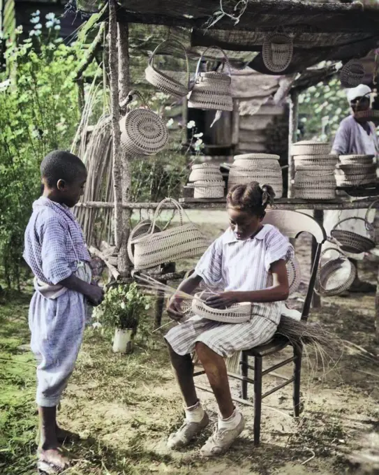 1930s AFRICAN AMERICAN GULLAH BOY STANDING WATCHING GIRL WEAVING BASKET AT STAND SEVERAL BASKETS FOR SALE SOUTH CAROLINA USA