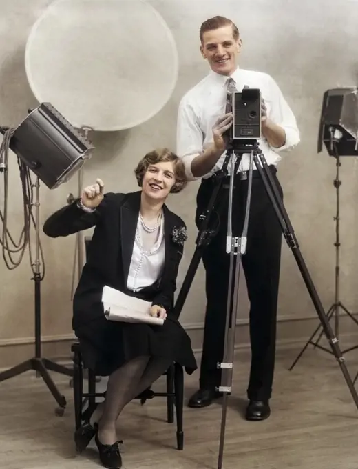 1920s PHOTOGRAPHER STANDING BEHIND MOVIE CAMERA IN STUDIO WITH ASSISTANT SITTING BESIDE HOLDING SCRIPT BOTH LOOKING AT CAMERA