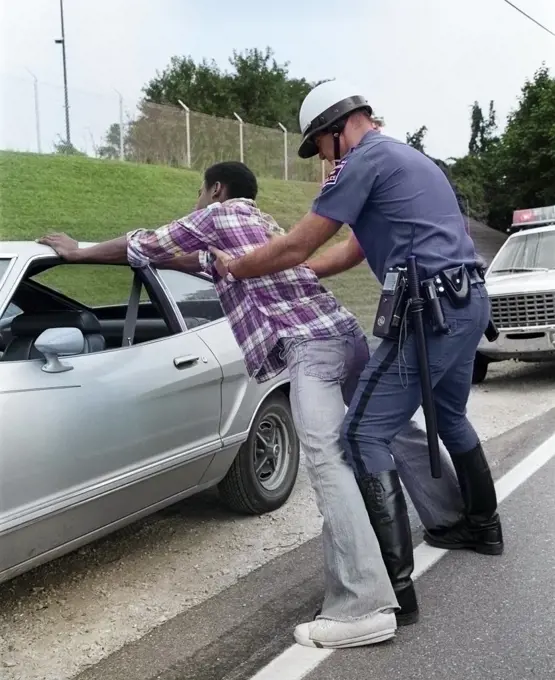 1970s CAUCASIAN POLICEMAN FRISKING SEARCHING AFRICAN AMERICAN TEENAGE MAN SPREAD EAGLE AGAINST ROADSIDE STOPPED MOTOR VEHICLE