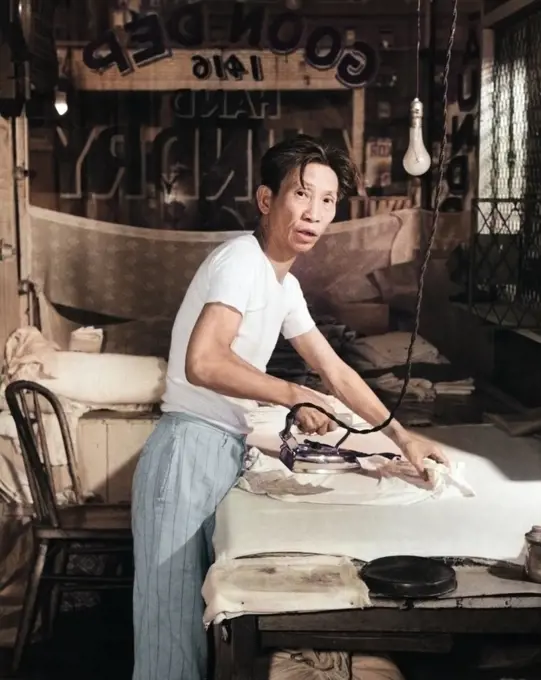 1920s CHINESE LAUNDRY MAN LOOKING AT CAMERA WORKING HARD IRONING CLOTHES BY HAND ON FLAT BOARD