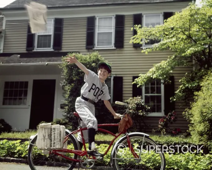1960s BOY IN BASEBALL UNIFORM RIDING BICYCLE DELIVERING NEWSPAPERS