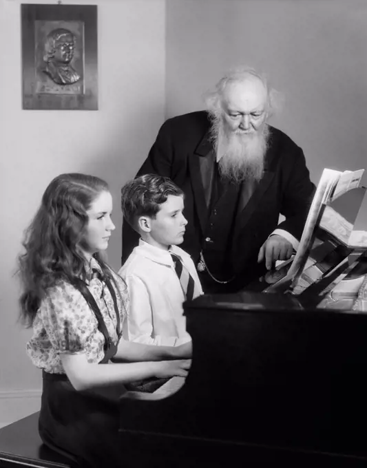 1930s 1940s BOY GIRL SEATED AT PIANO WITH ELDERLY MAN TEACHER WATCHING THEM PLAY