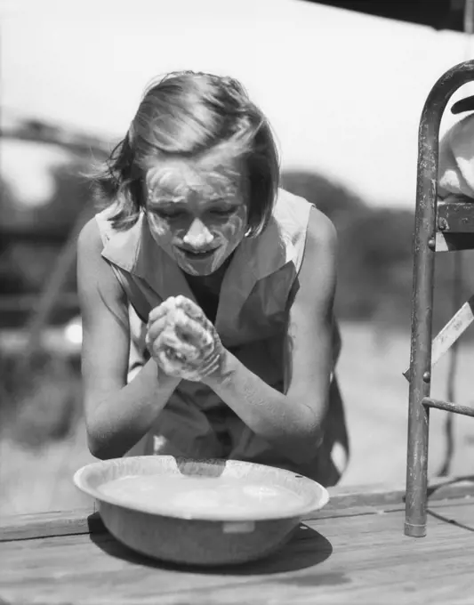 1930s YOUNG GIRL LEANING OVER BOWL WASHING HANDS AND FACE WITH SOAP AT SUMMER CAMP OUTDOORS 