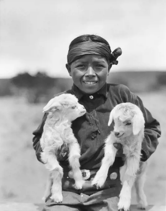 1930s SMILING NATIVE AMERICAN NAVAJO BOY LOOKING AT CAMERA HOLDING TWO BABY LAMBS NEW MEXICO USA