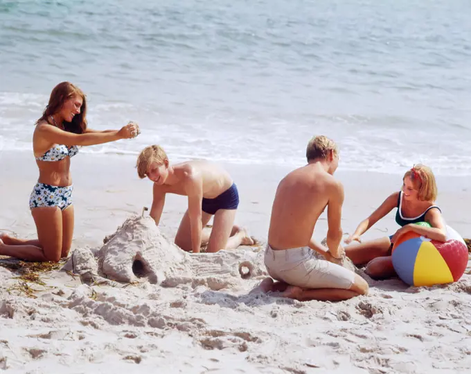 1960s 1970s TWO TEENAGE COUPLES ON BEACH BUILDING SANDCASTLE AND PLAYING WITH BALL