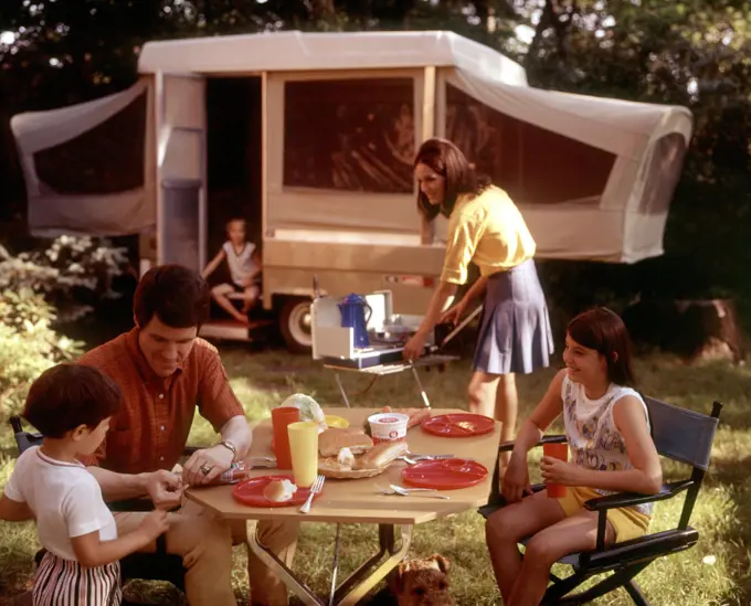 1970s FAMILY CAMPING VACATION CAMPSITE MOTHER COOKING ON CAMP STOVE FATHER AND KIDS AT TABLE EATING
