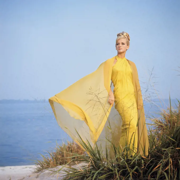 1960s 1970s BLOND WOMAN IN YELLOW GOWN STANDING IN BREEZE ON BEACH SAND DUNE 