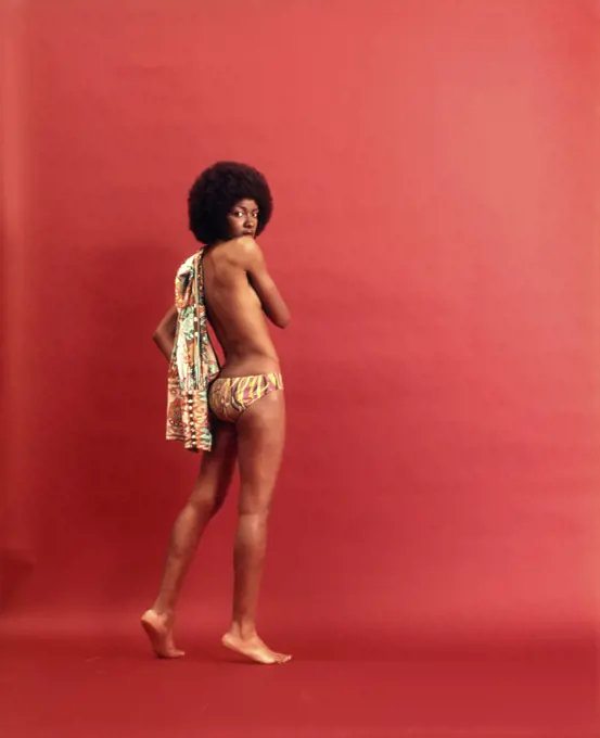 1970s AFRICAN AMERICAN WOMAN WITH AFRO HAIR STYLE WEARING TOPLESS BATHING SUIT STANDING TURNED AWAY LOOKING AT CAMERA