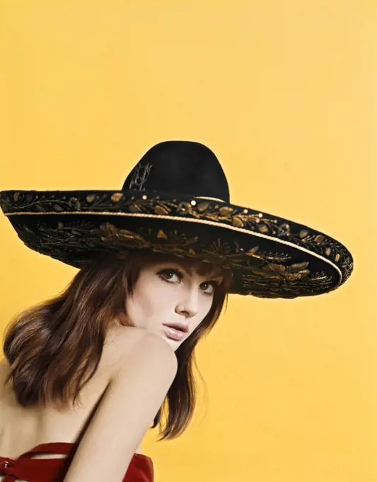 1970'S PORTRAIT ATTRACTIVE SULTRY BRUNETTE WOMAN WEARING GOLD EMBROIDERED BLACK MEXICAN SOMBRERO LOOKING AT CAMERA OVER SHOULDER
