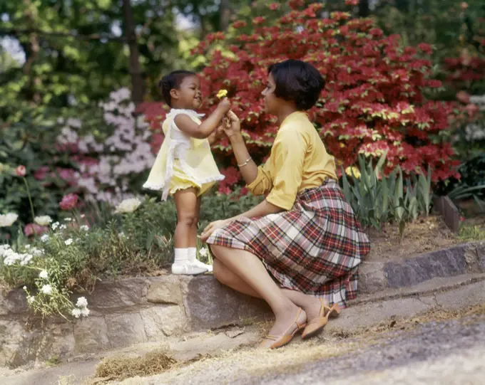 1970s SMILING AFRICAN AMERICAN WOMAN AND LITTLE GIRL OUTDOORS NEAR SPRING FLOWERS DAUGHTER GIVING YELLOW FLOWER TO MOTHER