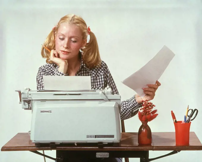 1970s YOUNG WOMAN BLONDE HAIR AT ELECTRIC HERMES TYPEWRITER THOUGHTFUL PENSIVE STUDENT TYPING WEARING HAIR IN PIGTAILS 