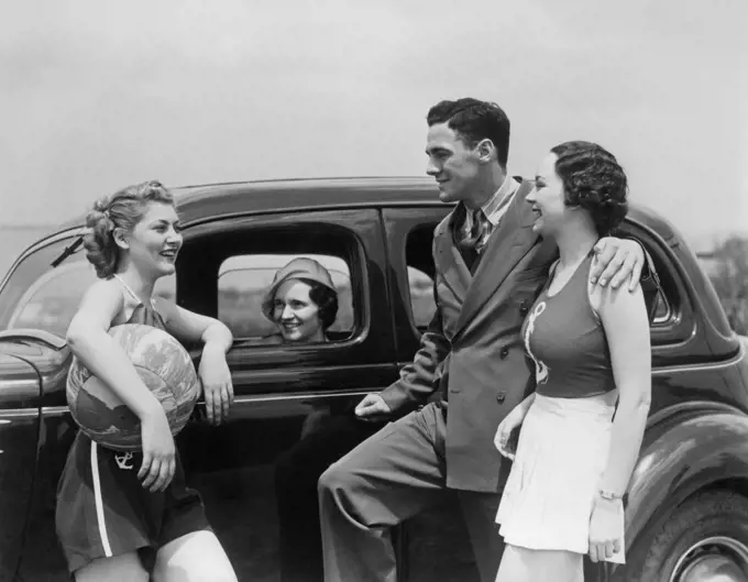 1930s TWO WOMEN IN NAUTICAL THEME CLOTHES AND MAN IN A SUIT TALKING STANDING BY THIRD WOMAN IN PARKED AUTOMOBILE AT SEASHORE