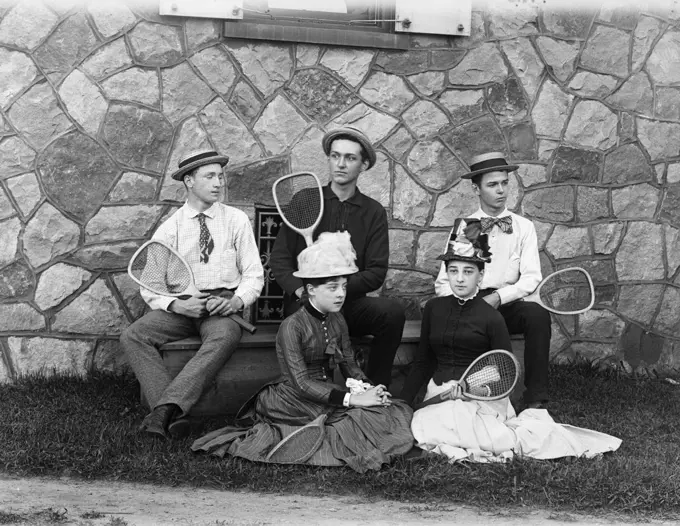1890s 1900s THREE TEENAGED BOYS SITTING ON BENCH TWO TEENAGED GIRLS SITTING ON GRASS ALL HOLDING TENNIS RACQUETS