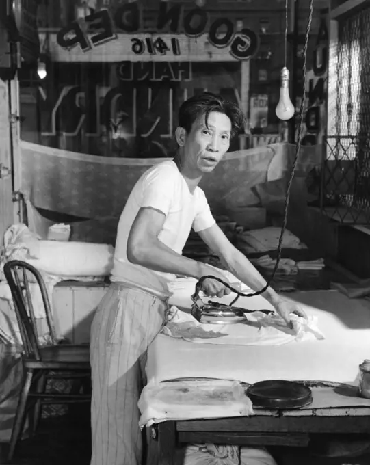 1920s CHINESE LAUNDRY MAN LOOKING AT CAMERA IRONING CLOTHES ON BOARD