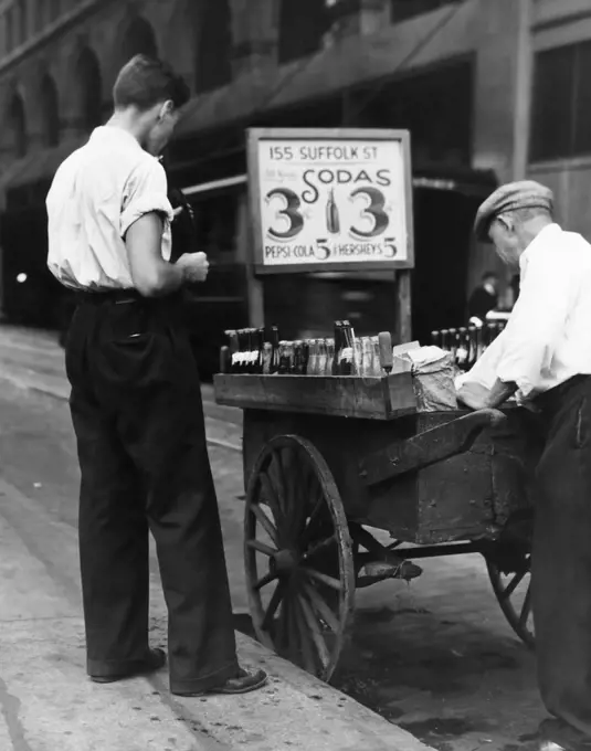 1930s YOUNG MAN CUSTOMER AND SIDEWALK STREET VENDER WITH HANDCART SELLING SODAS FOR 3 CENTS & 5 CENTS FOR PEPSI COLA NYC USA