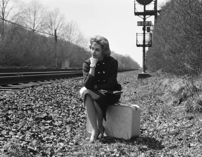 1950s 1960s WOMAN SAD WORRIED FACIAL EXPRESSION SITTING  ON SUITCASE BY SIDE RAILROAD TRACKS
