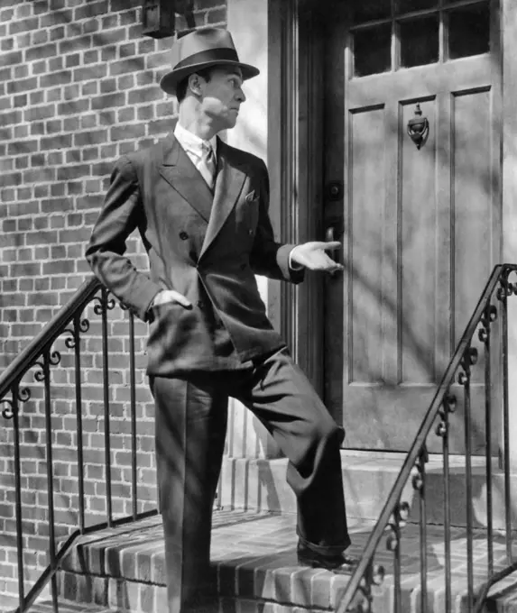 1950s MAN SUIT TIE HAT SALESMAN STANDING REJECTED AT FRONT DOOR ON FRONT STOOP OF BRICK BUILDING