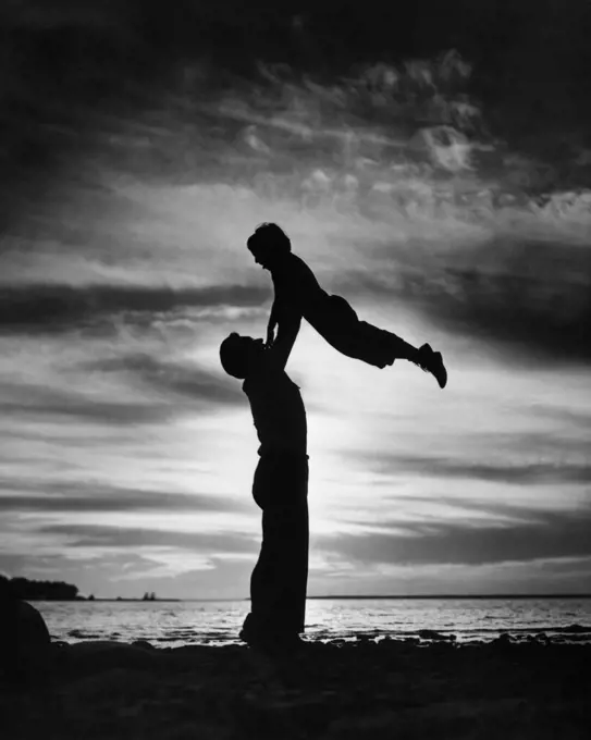 1940s 1950s ANONYMOUS SILHOUETTED MAN FATHER LIFTING CHILD SON IN AIR ON SHORE AT DUSK 