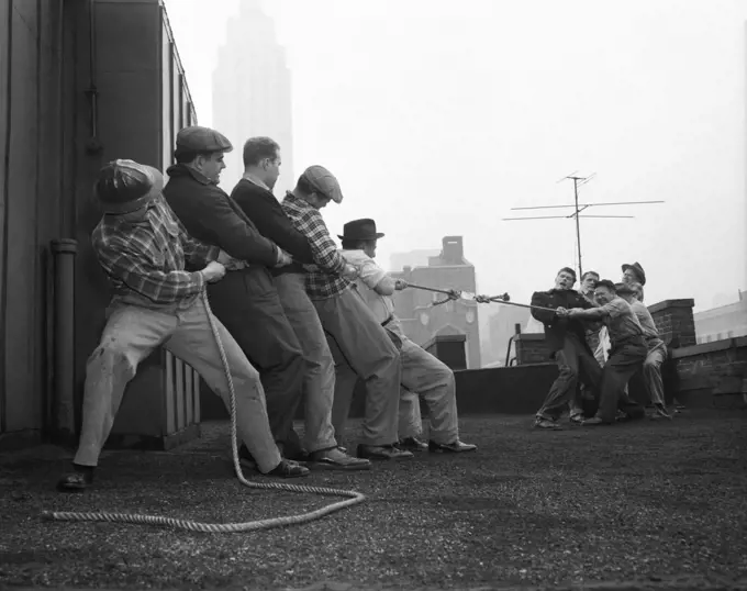 1950s TWO GROUPS OF FIVE MEN EACH PULLING ON TUG-OF-WAR ROPE ON BUILDING ROOFTOP TRYING TO PULL SOMETHING APART