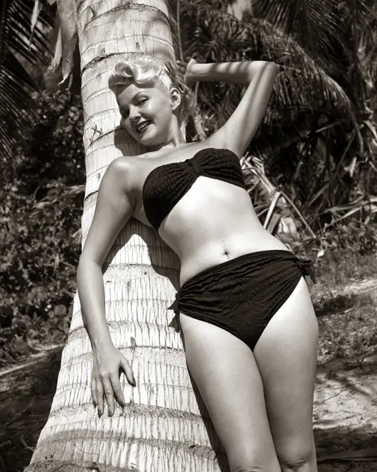 1950s SEXY SMILING BLONDE WOMAN IN BLACK TWO PIECE BIKINI BATHING SUIT SWIM WEAR LEANING AGAINST PALM TREE LOOKING AT CAMERA