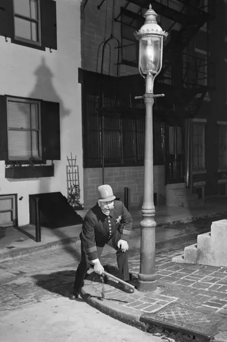 1900s MAN KEYSTONE COP UNIFORM STANDING ON STREET CORNER UNDER GAS LIGHT RAISING ALARM STRIKING SIDEWALK WITH BILLY CLUB