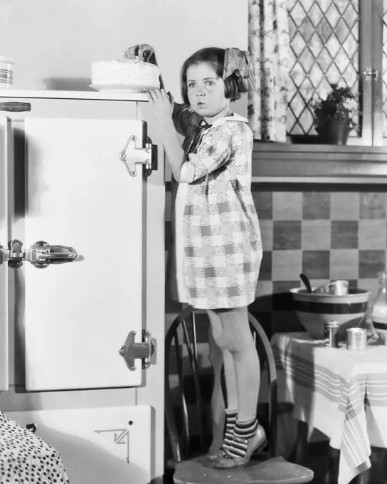 1920s 1930s LITTLE GIRL STANDING ON KITCHEN CHAIR TO REACH CAKE ON TOP OF ICE BOX 