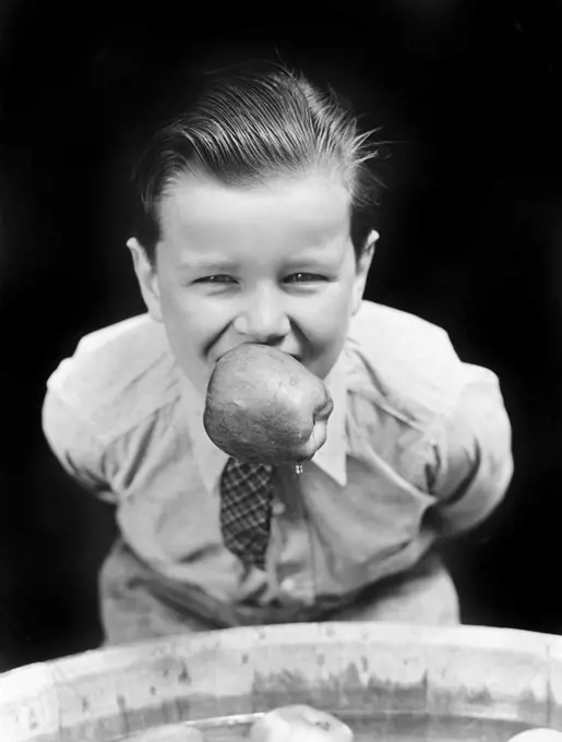 1930s BOY BOBBING FOR APPLES WITH AN APPLE IN HIS MOUTH LOOKING AT CAMERA