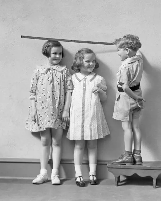 1930s BOY STANDING ON A STOOL MEASURING THE HEIGHT OF TWO GIRLS WITH YARD STICK