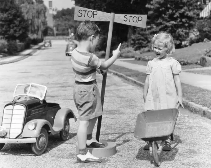 1930s 1940s BOY PLAYING TRAFFIC COP POINTING TO STOP SIGN HALTING GIRL WITH WHEELBARROW