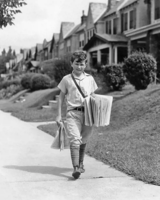 1930s BOY NEWSBOY LOOKING AT CAMERA DELIVERING NEWSPAPERS WALKING ALONG SUBURBAN STREET OF ROW HOUSES