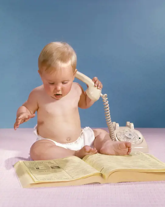 1960s BLOND BABY HOLDING TELEPHONE LOOKING AT PHONEBOOK YELLOW PAGES