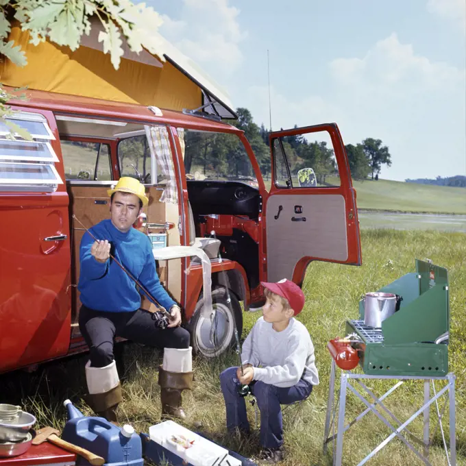 1970s MAN AND BOY CAMPING SON SITTING BY PORTABLE STOVE FATHER SITTING BY CAMPER WITH FISHING RODS AND GEAR           