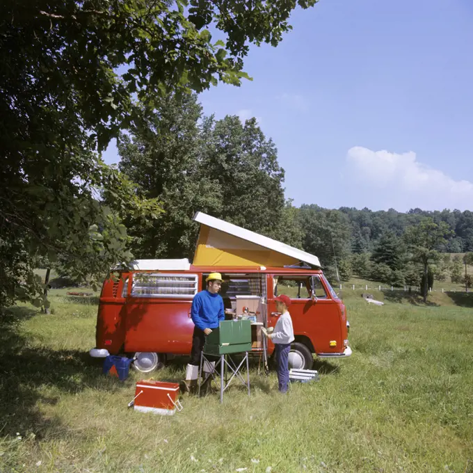 1970s FATHER AND SON COOKING AT CAMPSITE BESIDE RED POP TOP VW CARAVAN CAMPER