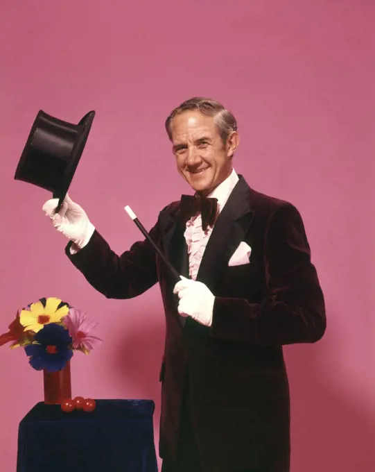 1970s MAN MAGICIAN LOOKING AT CAMERA TIPPING TOP HAT WITH WHITE GLOVES BOW TIE MAGIC WAND FLOWERS 
