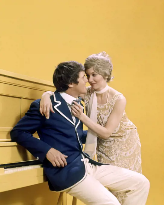 1970s COUPLE MAN WOMAN WEARING 1920s CLOTHES LEANING ON YELLOW UPRIGHT PIANO 