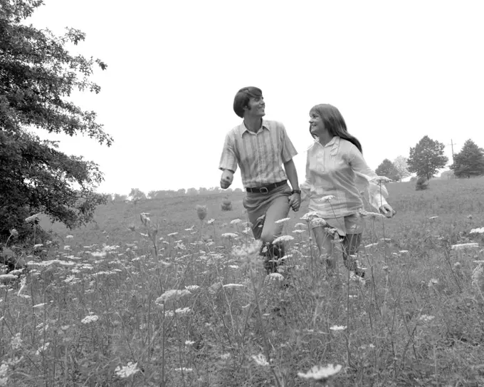 1970s COUPLE HOLDING HANDS WALKING IN HIGH FIELD WITH QUEEN ANNE'S LACE