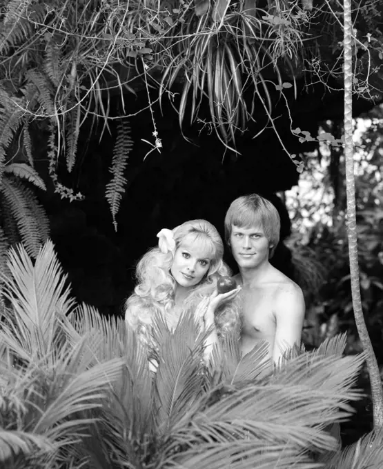 1970s BLONDE COUPLE POSED AS ADAM AND EVE LOOKING AT CAMERA STANDING NAKED BEHIND TROPICAL FOLIAGE EVE HOLDING APPLE