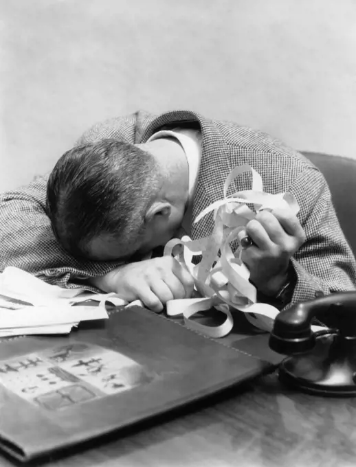 1930s 1940s MAN WITH HEAD DOWN ON DESK AND STOCK TICKER TAPE IN HAND 