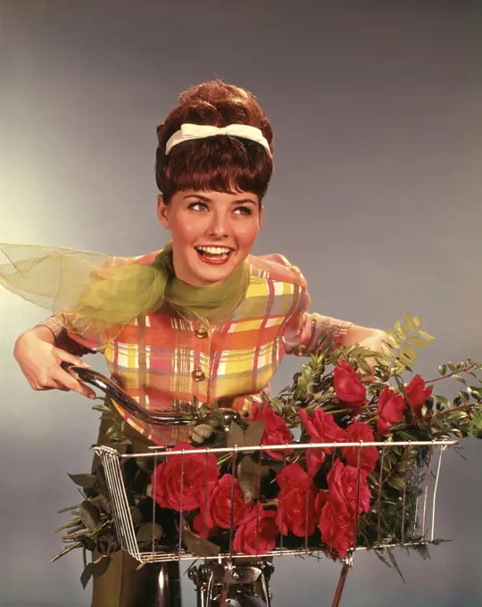 1960s SMILING TEEN GIRL WITH TEASED BRUNETTE BOUFFANT HAIRDO RIDING BICYCLE WITH RED ROSES BOUQUET IN BASKET