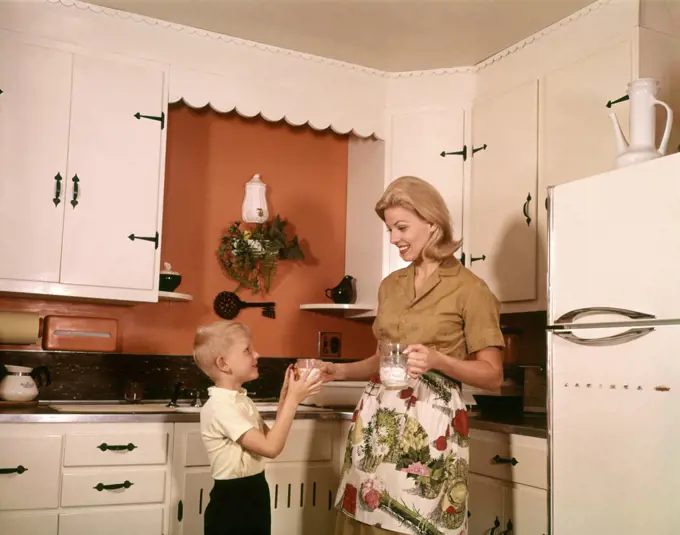 1970s BLONDE MOTHER GIVING SON GLASS MILK IN KITCHEN  