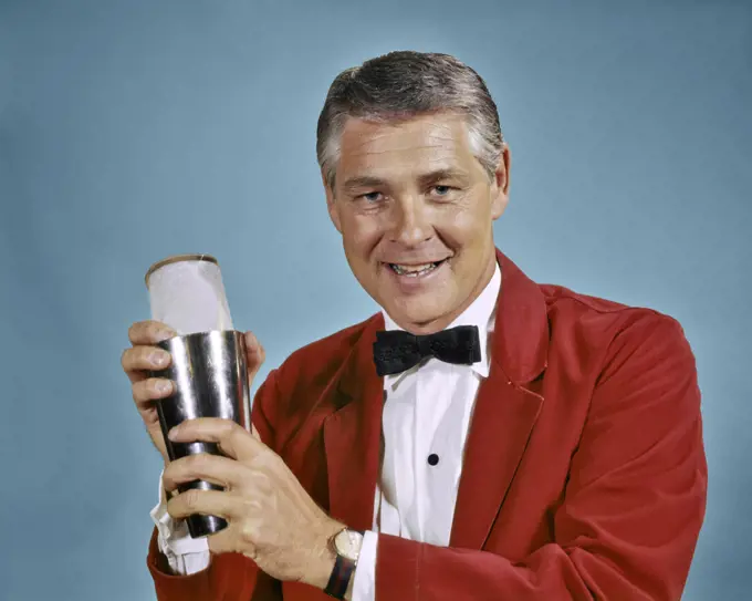 1960s 1970s SMILING MAN BARTENDER IN RED JACKET LOOKING AT CAMERA HOLDING COCKTAIL SHAKER MAKING MIXED DRINK