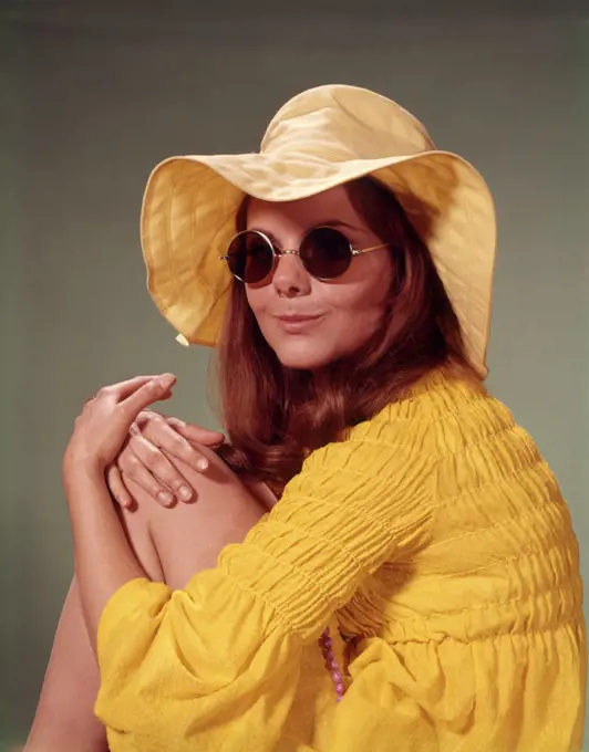 1960s PORTRAIT SMILING BRUNETTE TEENAGE WOMAN IN YELLOW FLOPPY BRIM HAT SMOCKED SHIRT ROUND LENS SUNGLASSES LOOKING AT CAMERA