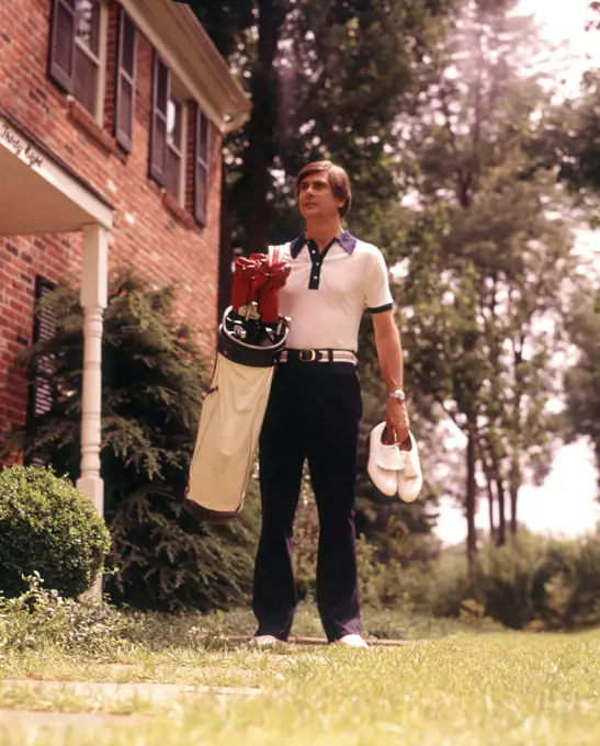 1970s MAN CASUALLY DRESSED CARRYING GOLF CLUBS AND SHOES LEAVING FRONT OF SUBURBAN BRICK HOUSE