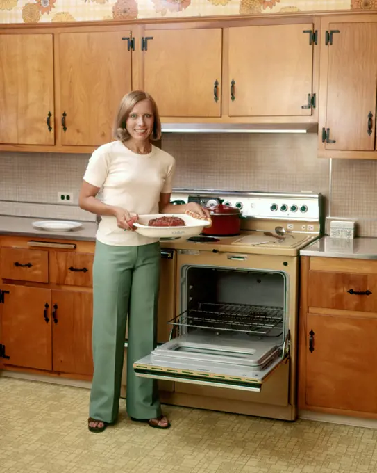 1970s SMILING BLOND WOMAN HOUSEWIFE LOOKING AT CAMERA HOLDING BAKED HAM STANDING BY STOVE OPEN OVEN DOOR