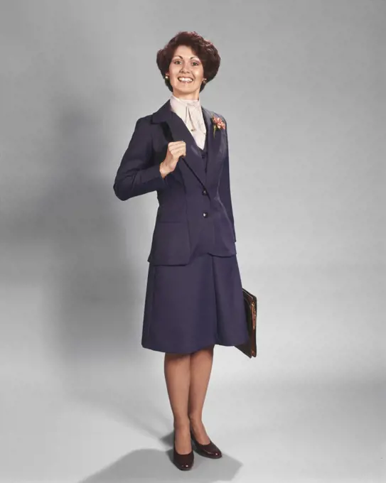 1970s CONFIDENT HAPPY SMILING BUSINESSWOMAN STANDING WEARING BLUE BUSINESS SUIT HOLDING BRIEFCASE LOOKING AT CAMERA