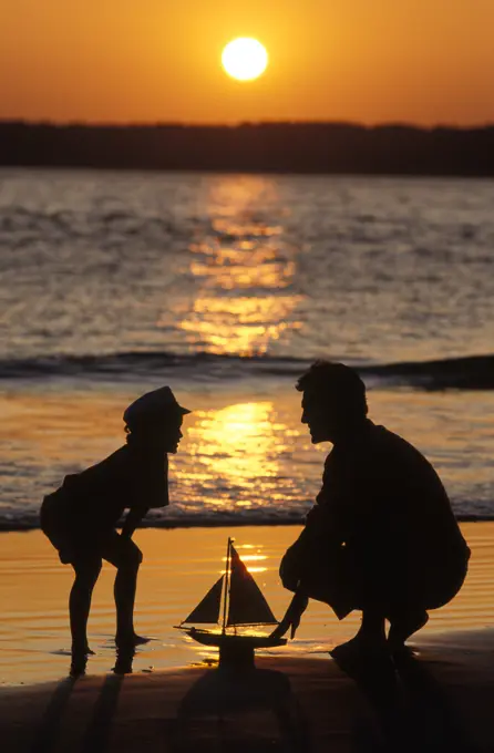 1990s ANONYMOUS FATHER AND SON PLAYING WITH TOY BOAT AT THE BEACH SILHOUETTED AGAINST SETTING SUN