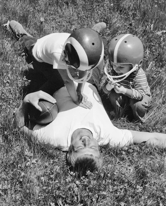 1970s MAN FATHER LYING IN GRASS HOLDING FOOTBALL AFTER BEING TACKLED BY SON AND DAUGHTER