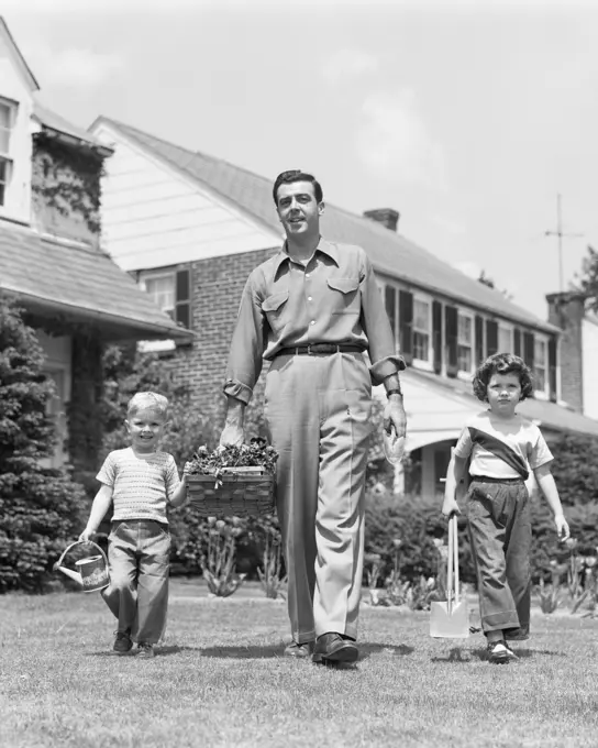 1950s FATHER WITH TWO CHILDREN DAUGHTER AND SON WALKING IN FRONT YARD CARRYING GARDENING TOOLS LOOKING AT CAMERA